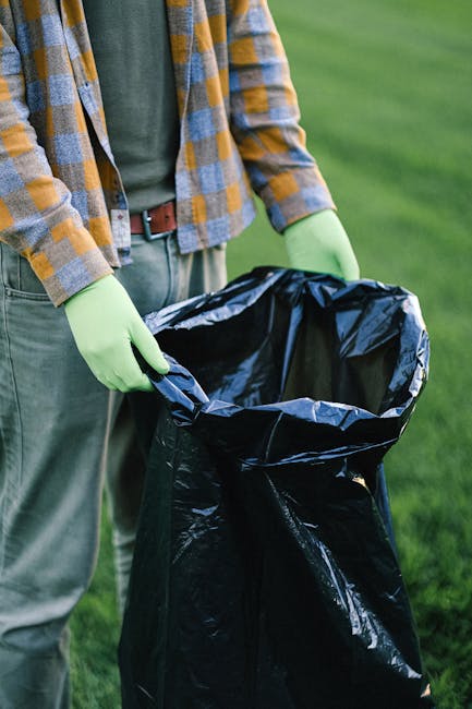 A person wearing a checkered shirt with shades of yellow, grey, and blue, along with green gloves, is holding open a large black rubbish bag made of plastic, with the bag's edges folded outward. The i